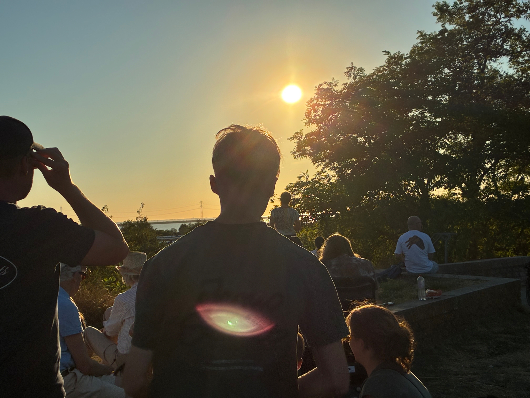 Auto-generated description: A group of people is gathered outdoors, watching the sun set over a natural landscape with trees.