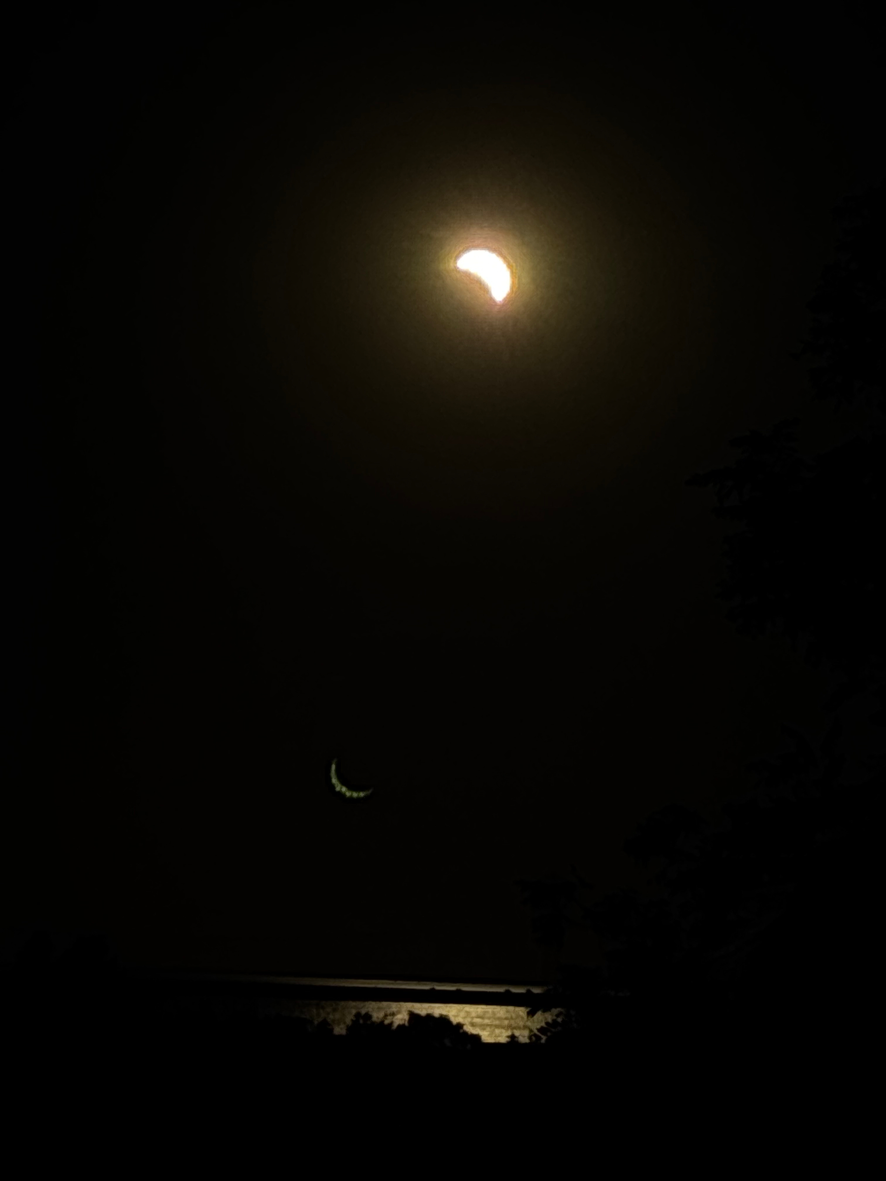 Auto-generated description: A bright crescent moon is prominently visible in the night sky with a faint, smaller crescent shape below it, and dark silhouettes of trees and a body of water reflecting moonlight at the bottom.