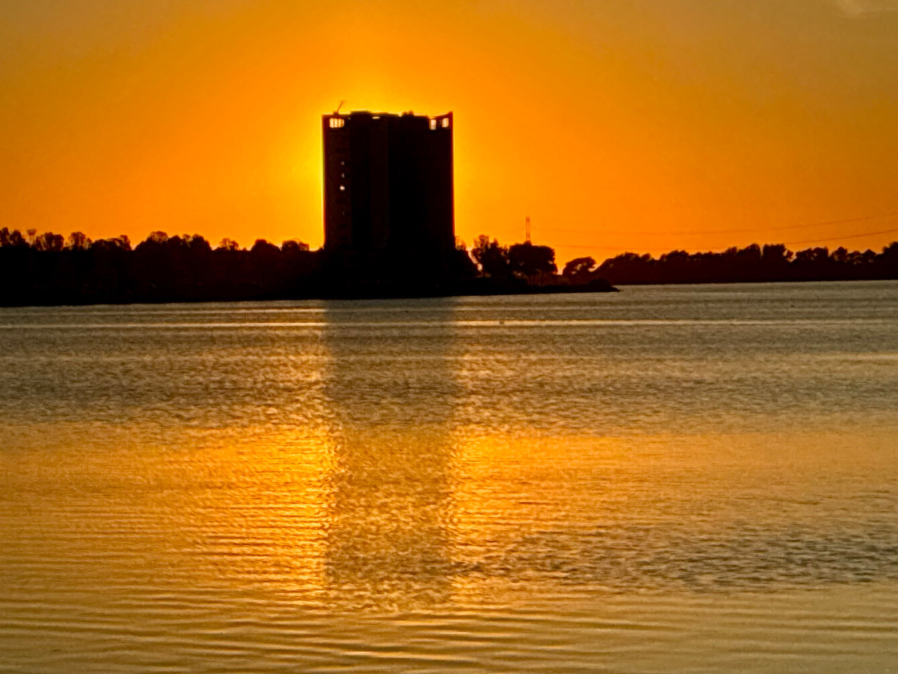 Auto-generated description: A tall building silhouetted against a vibrant orange sunset reflects on the calm water in the foreground.