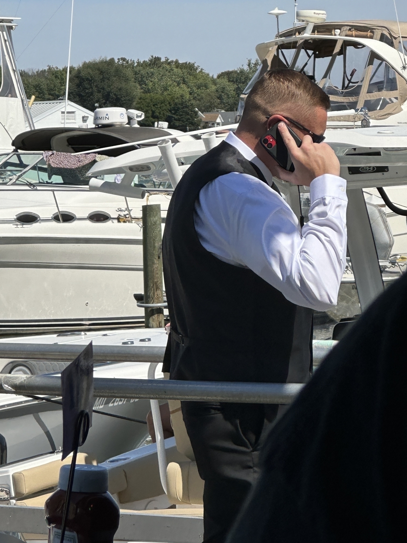 Man in black vest, white shirt, on cell phone surrounded by boats.  