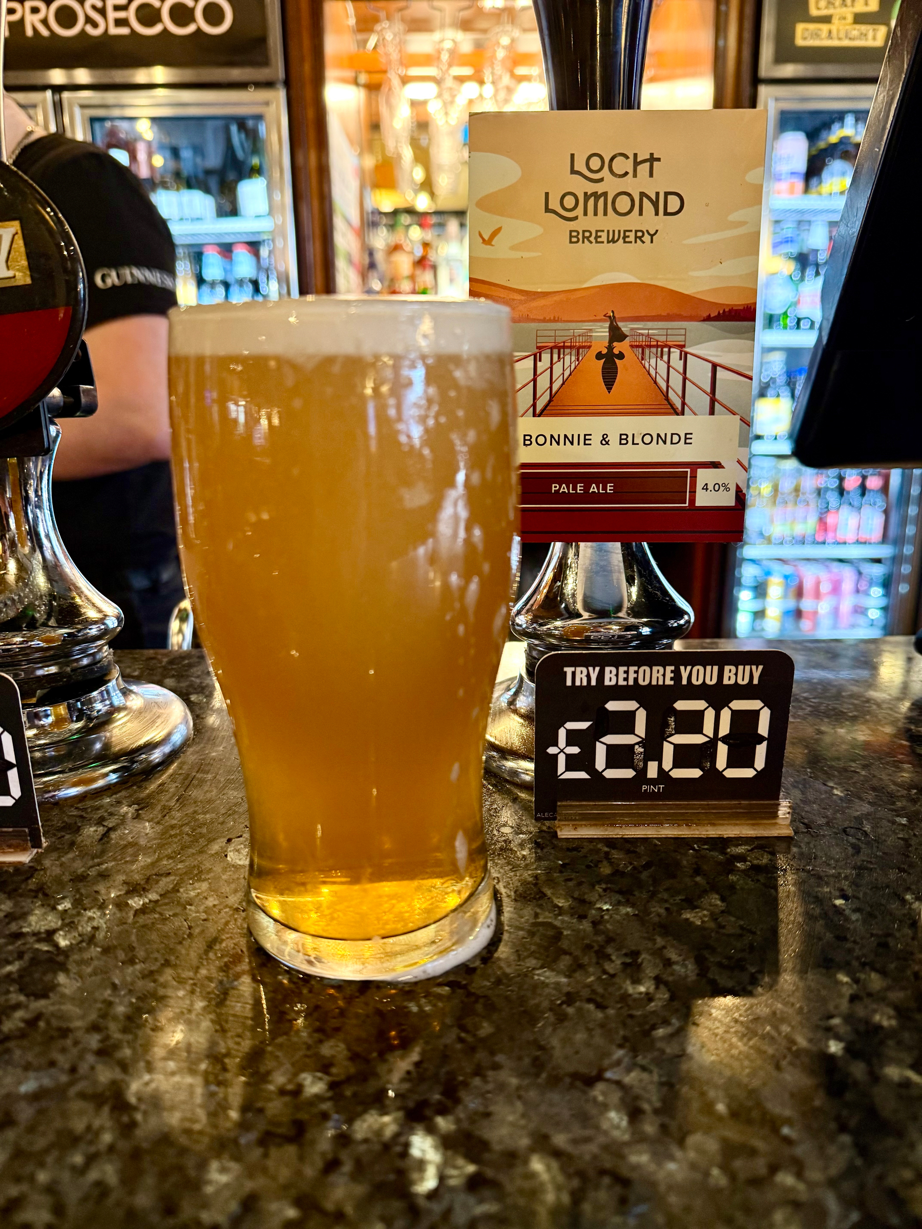 A pint of pale ale sits on a bar counter next to a digital price display and a promotional sign for Loch Lomond Brewery's Bonnie & Blonde.
