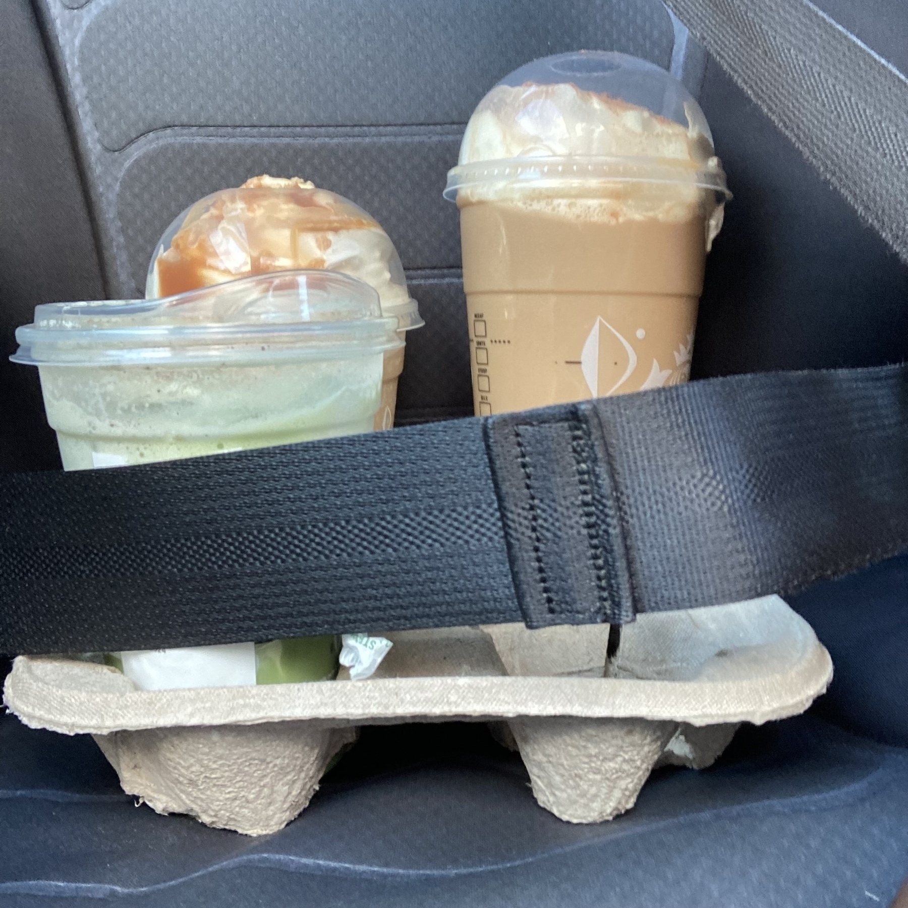 Two drinks, one caramel and one green tea, secured in a car seat with a seatbelt.