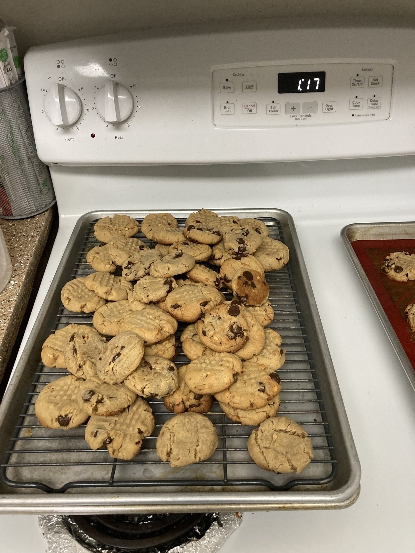 A batch of cookies, including ones with chocolate chips, are cooling on a wire rack placed over a baking sheet in a kitchen.