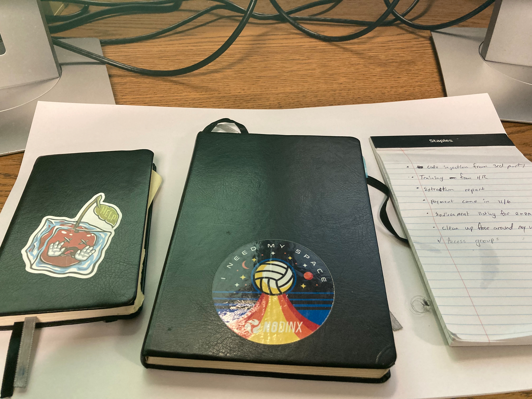 Two black notebooks, one with a sticker of an apple and the other with a space-themed sticker, are placed alongside a notepad with notes on a wooden desk.