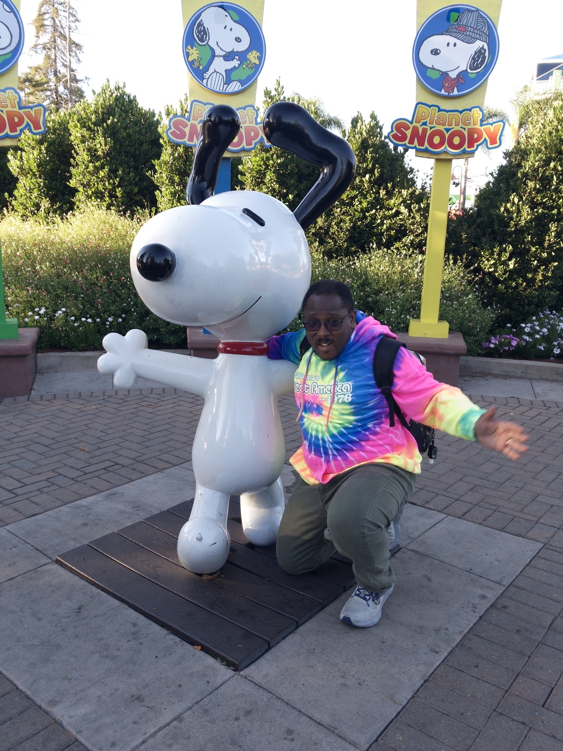 A person wearing a colorful tie-dye hoodie poses excitedly next to a statue of Snoopy.