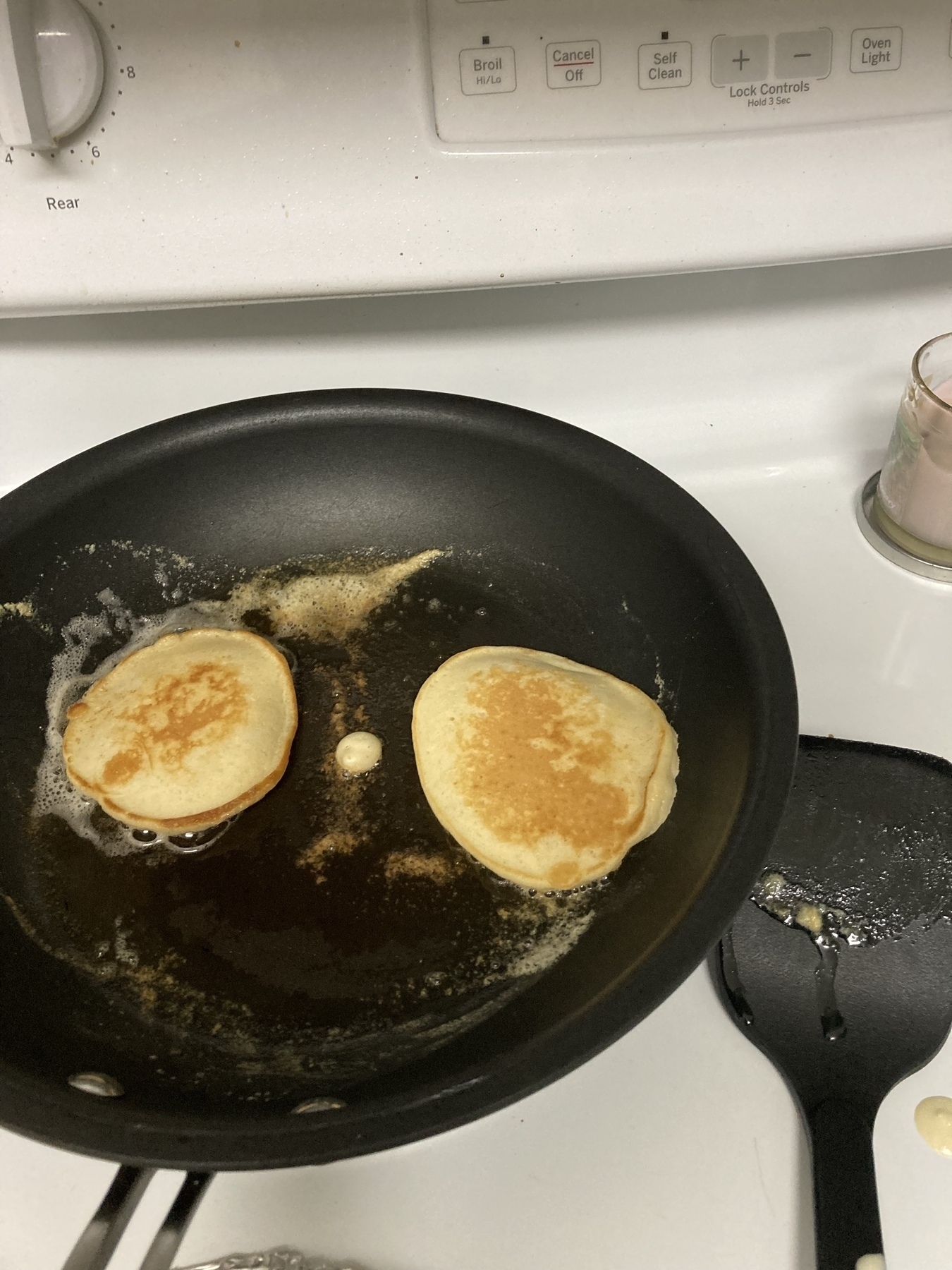 Two pancakes are cooking in a frying pan on a stovetop with a spatula nearby.