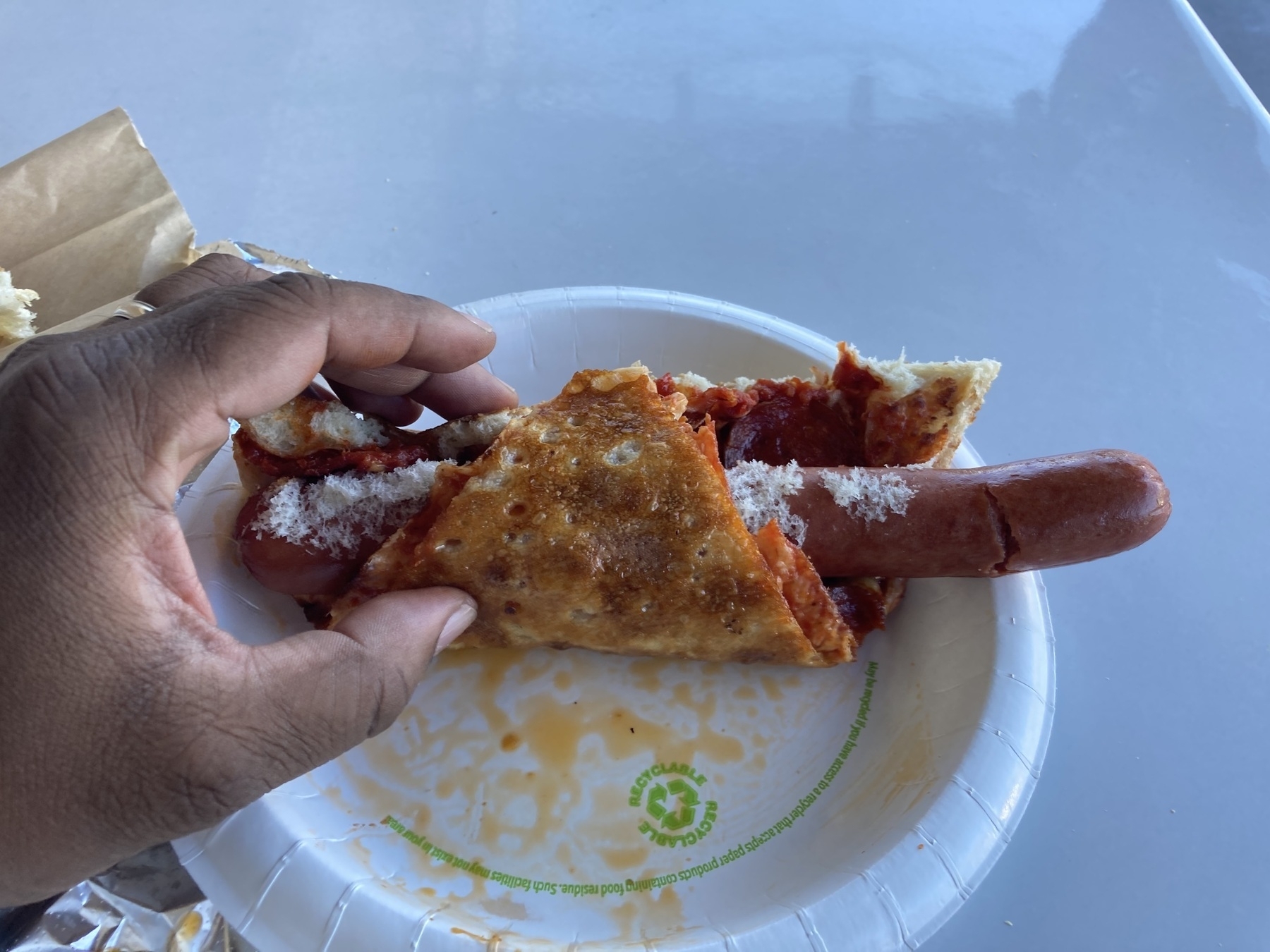 A hand is holding a hot dog wrapped in a thin, crispy crust on a paper plate with some sauce and cheese.