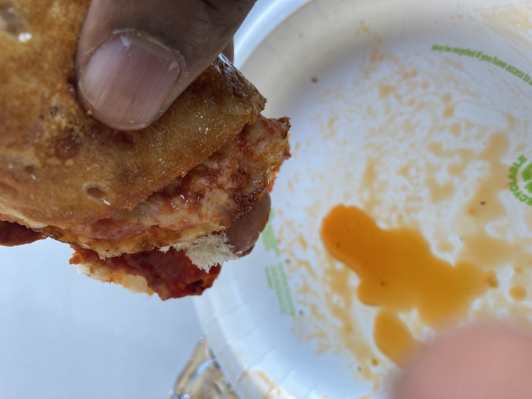 A hand holds a partially eaten sandwich filled with sauce, above a paper plate with some spilled sauce.