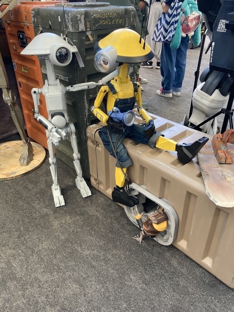 Two robotic figures, one white and one yellow, are sitting and interacting in a casual, public setting with people in the background.