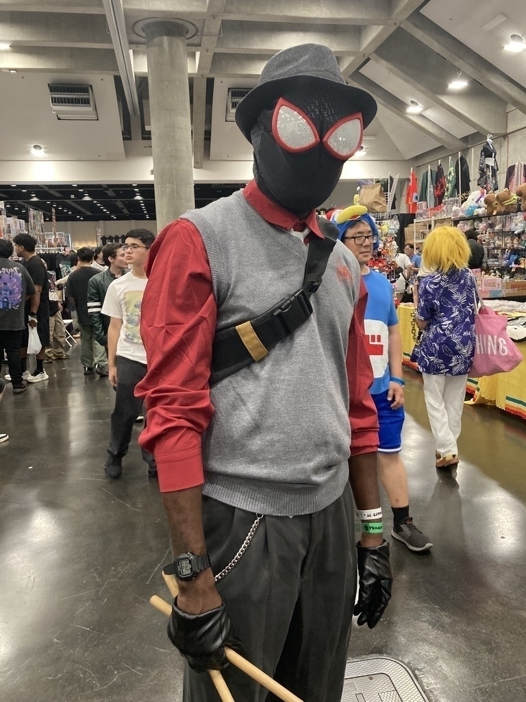 A person in a Spider-Man costume with a sweater vest, hat, and gloves stands holding drumsticks at a convention.