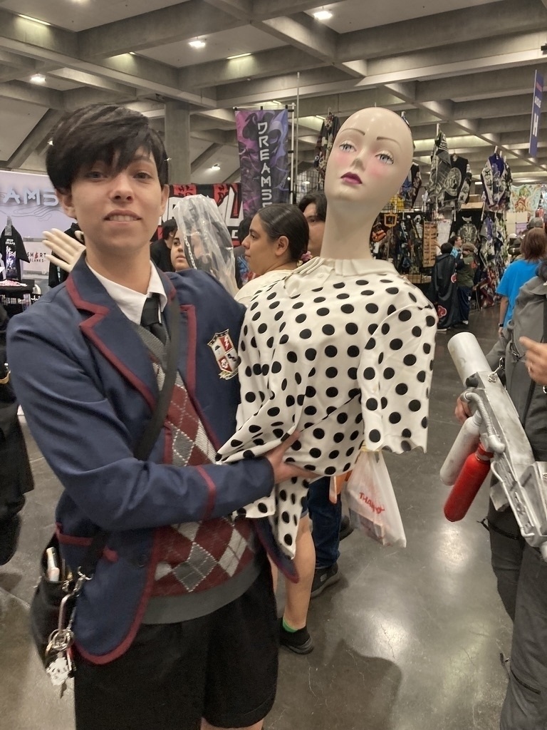 A person in a cosplay outfit holds a mannequin dressed in a polka dot outfit at a convention or event.