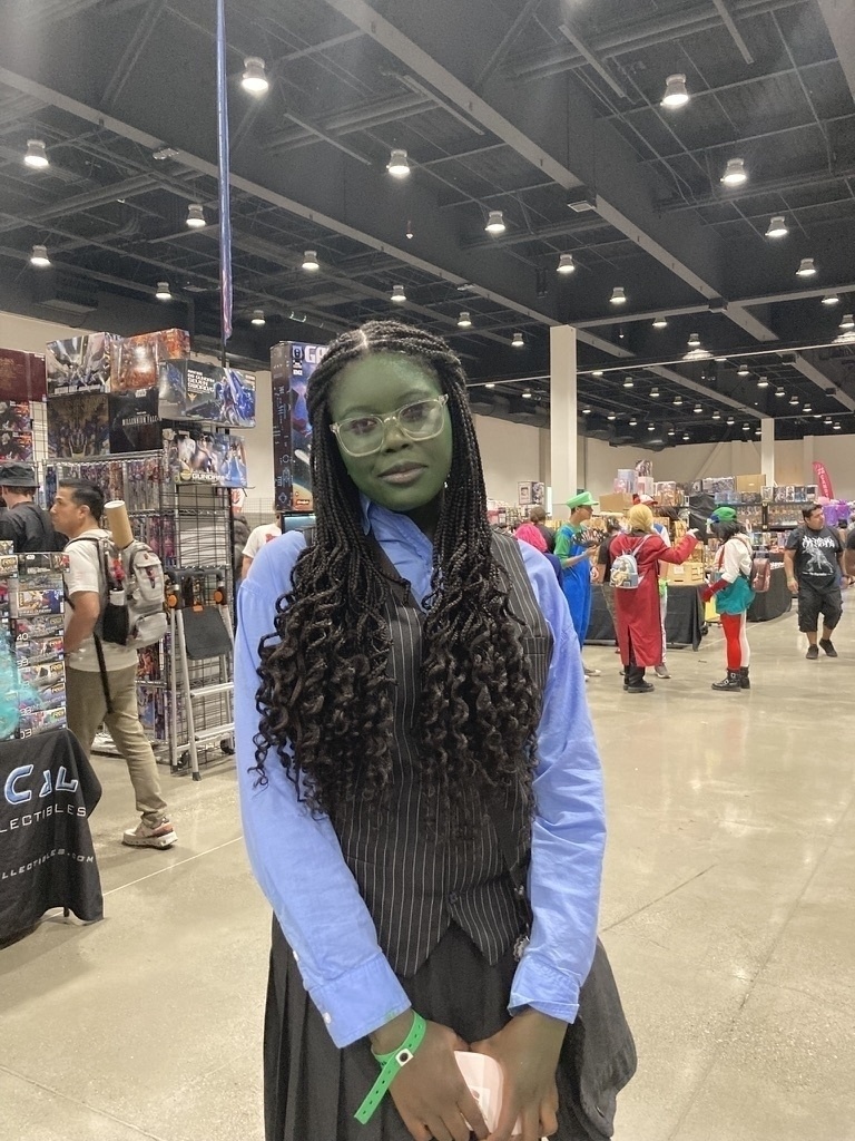 A person with green face paint dressed in a costume stands in a convention center surrounded by various booths and other attendees in colorful outfits.