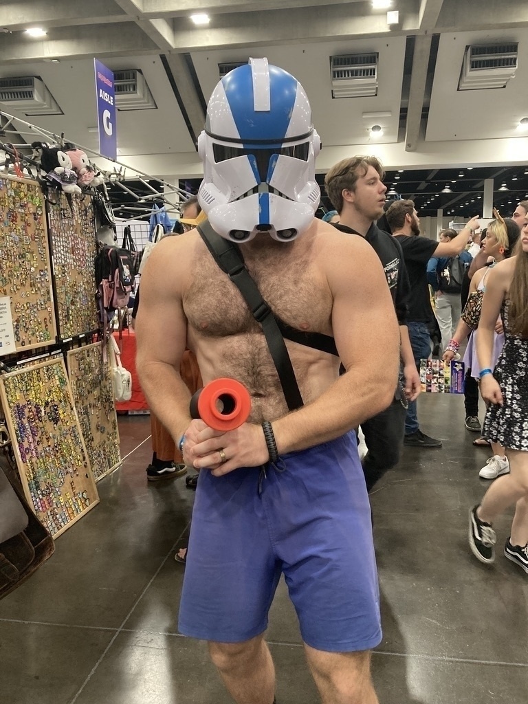 A person wearing a blue and white helmet with a blue bag and holding an item stands in a busy indoor event, surrounded by other attendees and vendor booths.