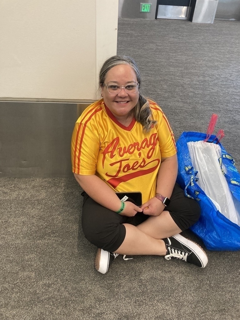 A person is sitting on the floor in a yellow Average Joe's jersey next to a large blue bag.