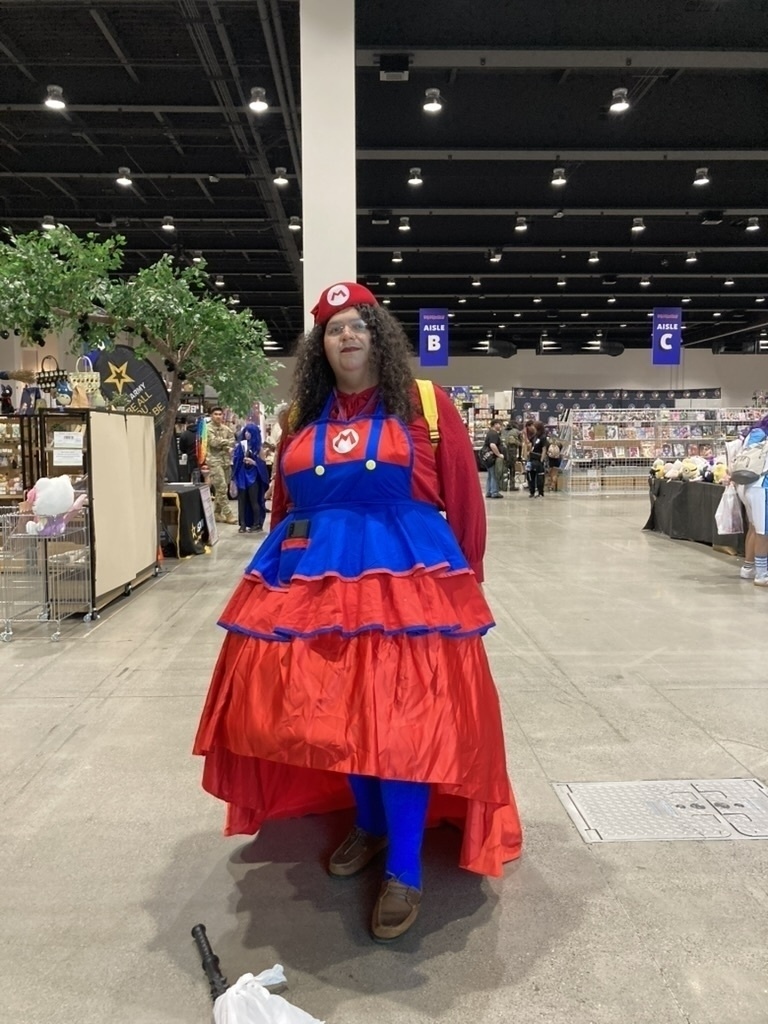 A person is dressed in a red and blue Mario-themed costume at a convention center, surrounded by various booths.