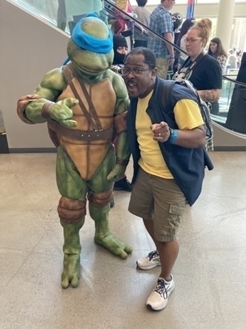 A person poses enthusiastically with someone dressed as a Teenage Mutant Ninja Turtle at an indoor event.