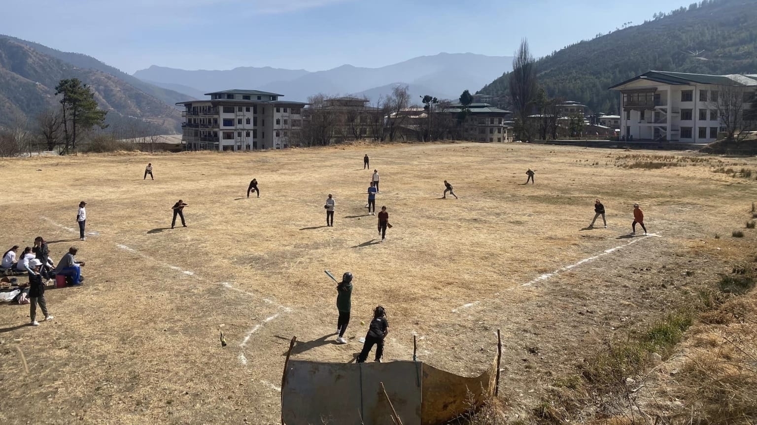 Kids playing baseball on a flattened field in Bhutan. The grass looks dry. . Buildings and mountains are visible in the background. 