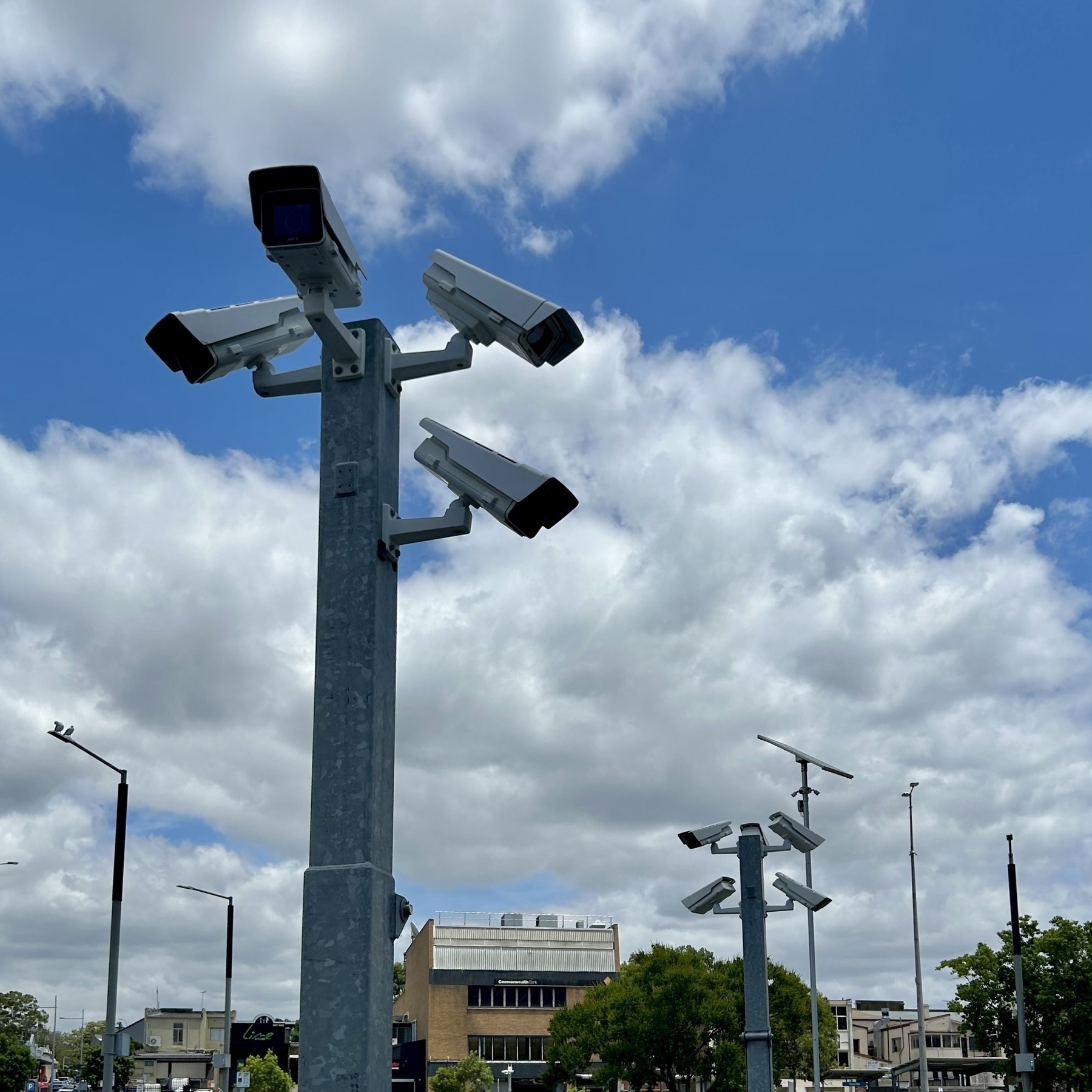 Two poles are visible, each with four surveillance cameras. Multiple light poles are visible in the distance, as are several buildings. 