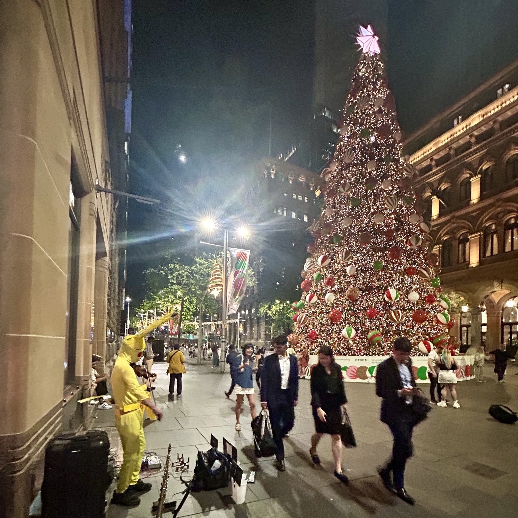 A person wearing a Pikachu costume is playing a violin on the left of the image. There is a giant Christmas tree on the right of the image. Several people are walking past, and looking at the violinist. ￼