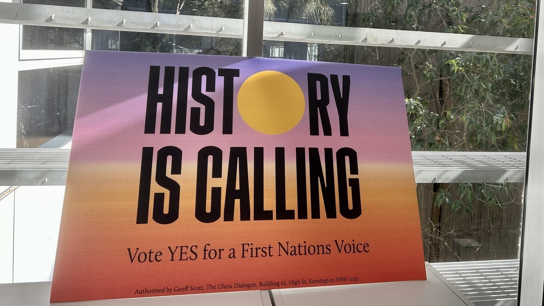 A corflute sign resting against a window. The text says: History is calling, vote Yes for a First Nations Voice