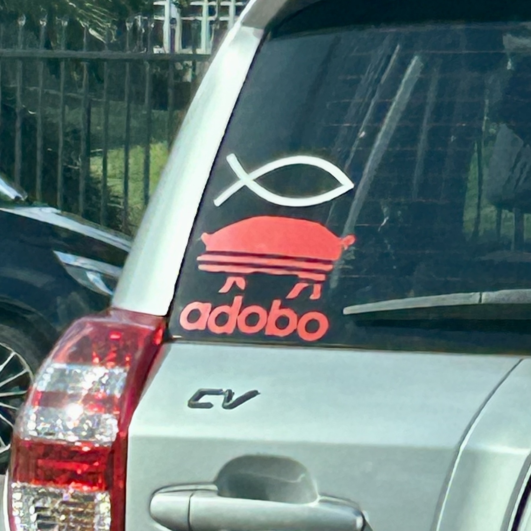 A car rear window. Below a Christian fish sticker is a pig with the word adobo on it, designed to look like an Adidas logo. It's referring to the Filipino dish pork adobo. 