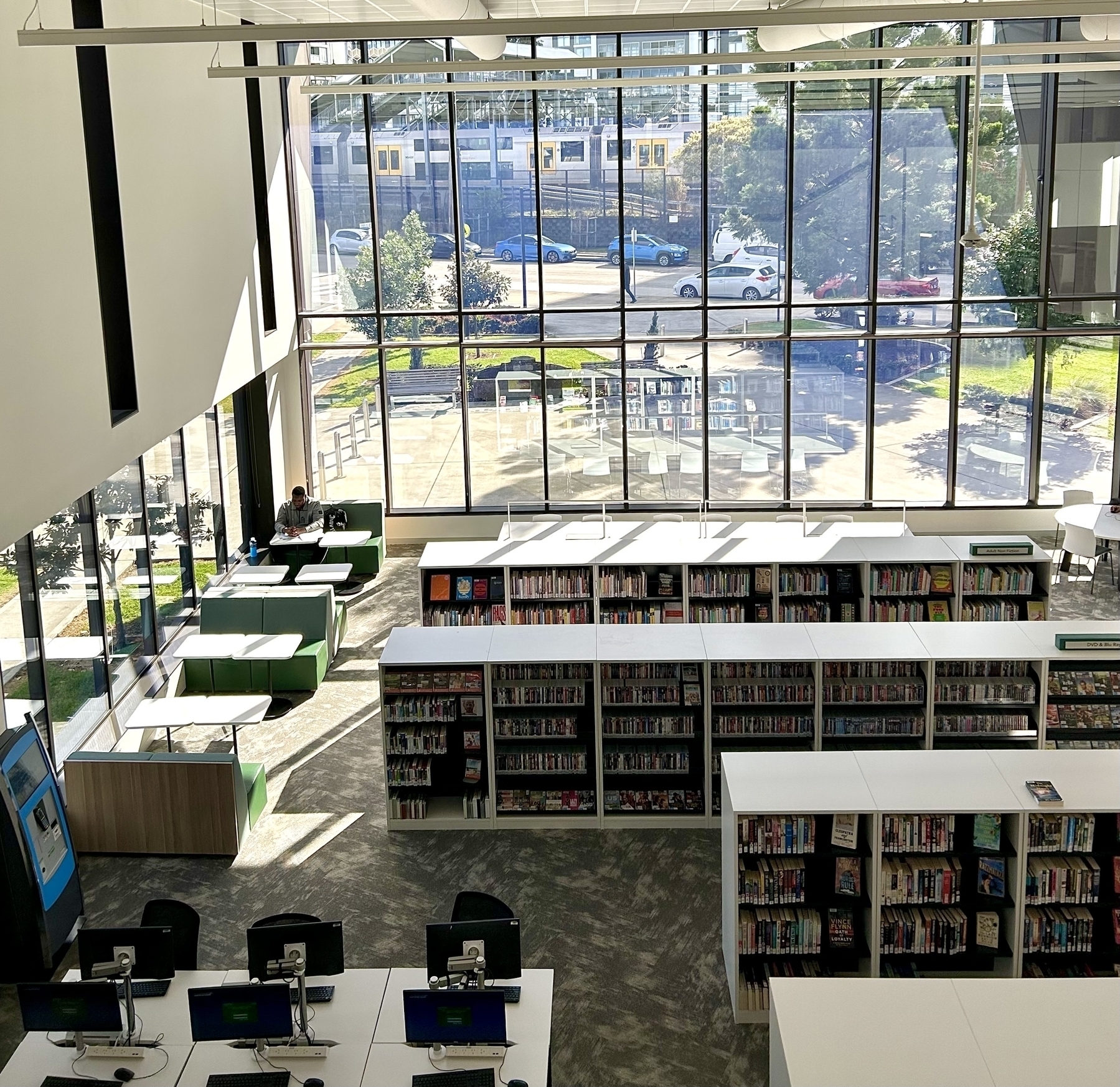 Looking down on a library from an elevated position. Rows of books and computers are visible in the foreground.  A large window looks out over some trees. A train is visible in the background. 