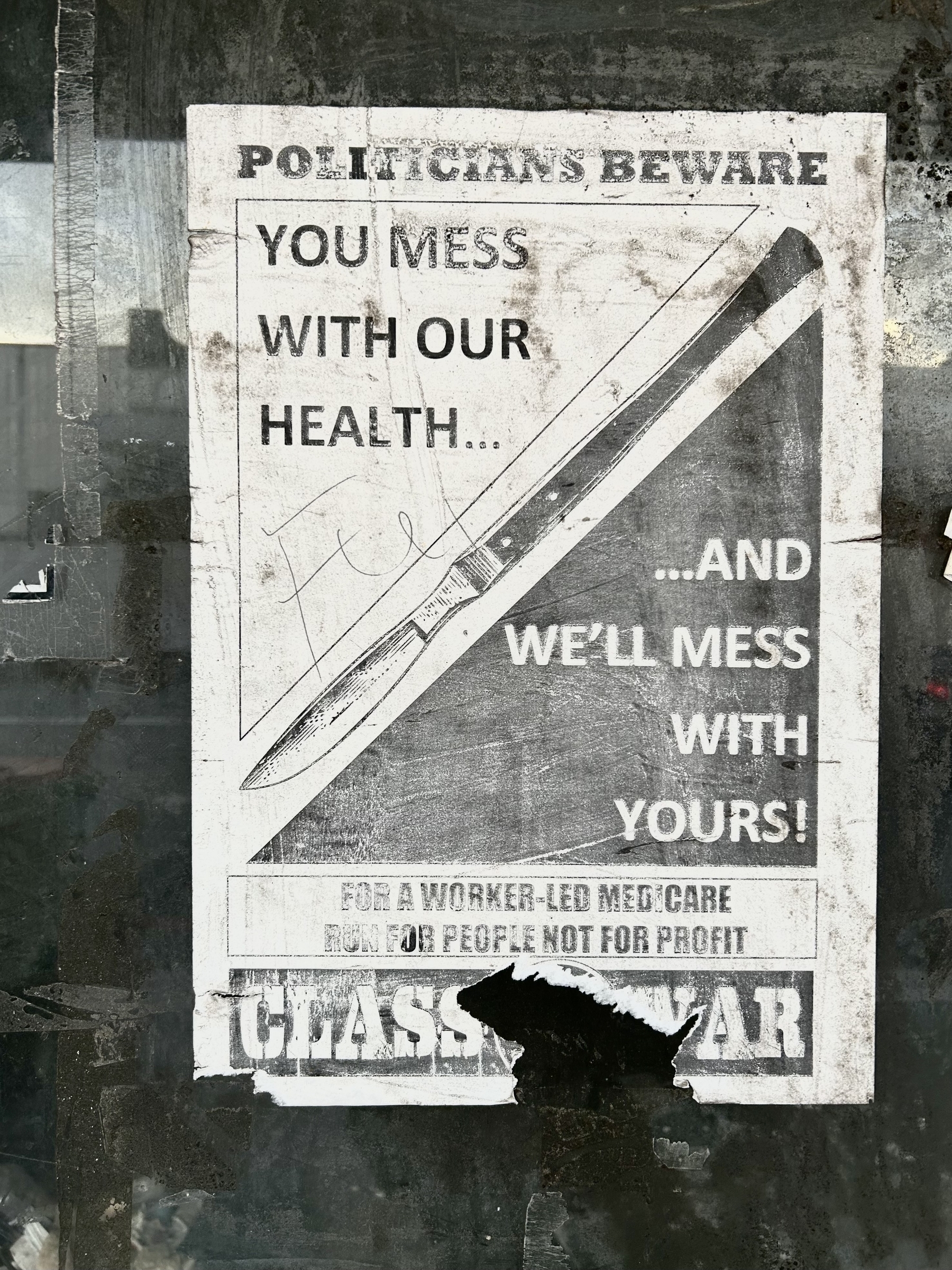 Paste up poster stuck to the window of a vacant shop. Text says: POLITICIANS BEWARE… YOU MESS WITH OUR HEALTH AND WE'LL MESS WITH YOURS.&10;Over a surgical scalpel. 