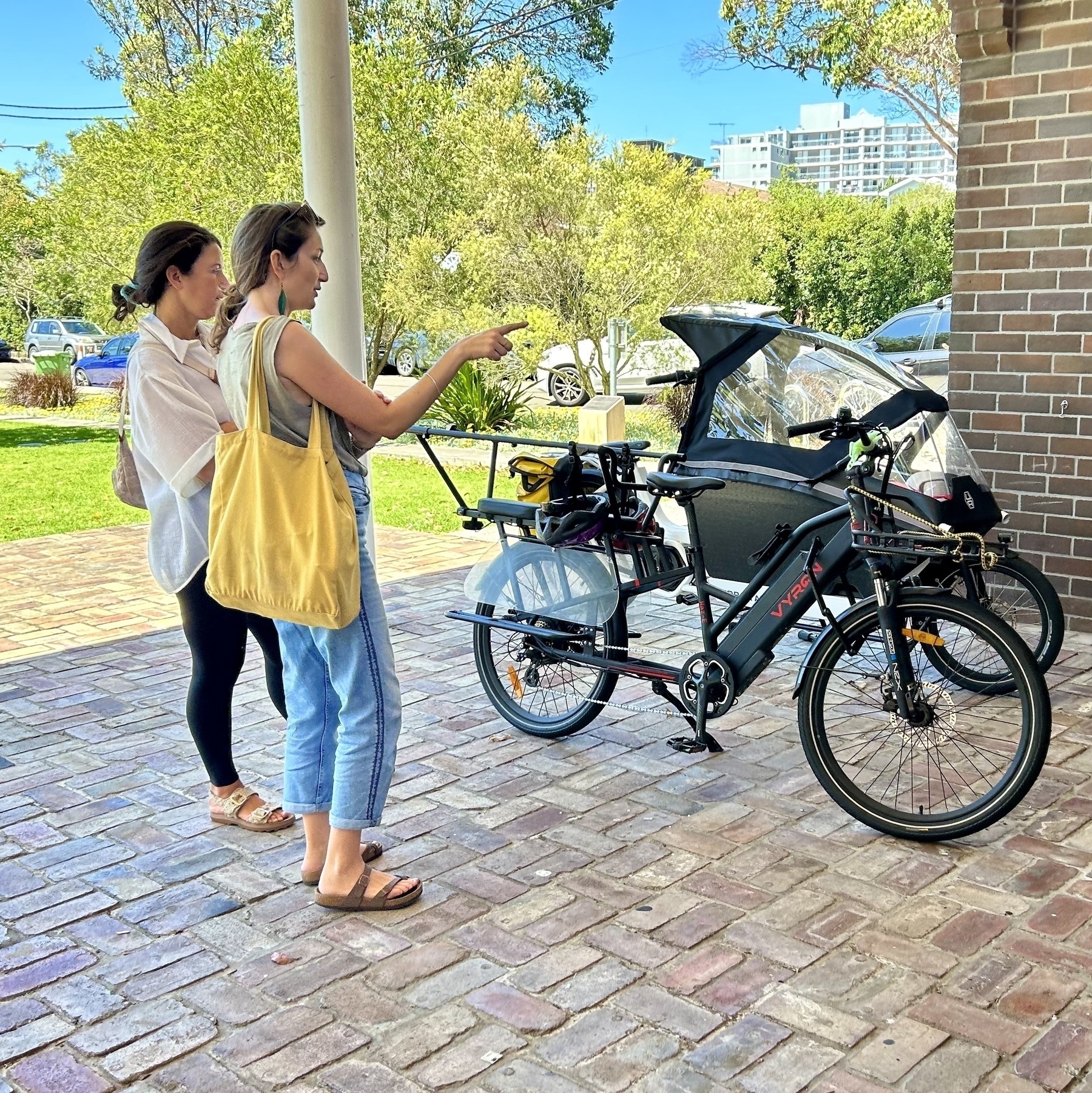 Two women looking at two parked e-bikes and gesturing at them while talking. One is a carrier bike and one is an enclosed cargo bike. 