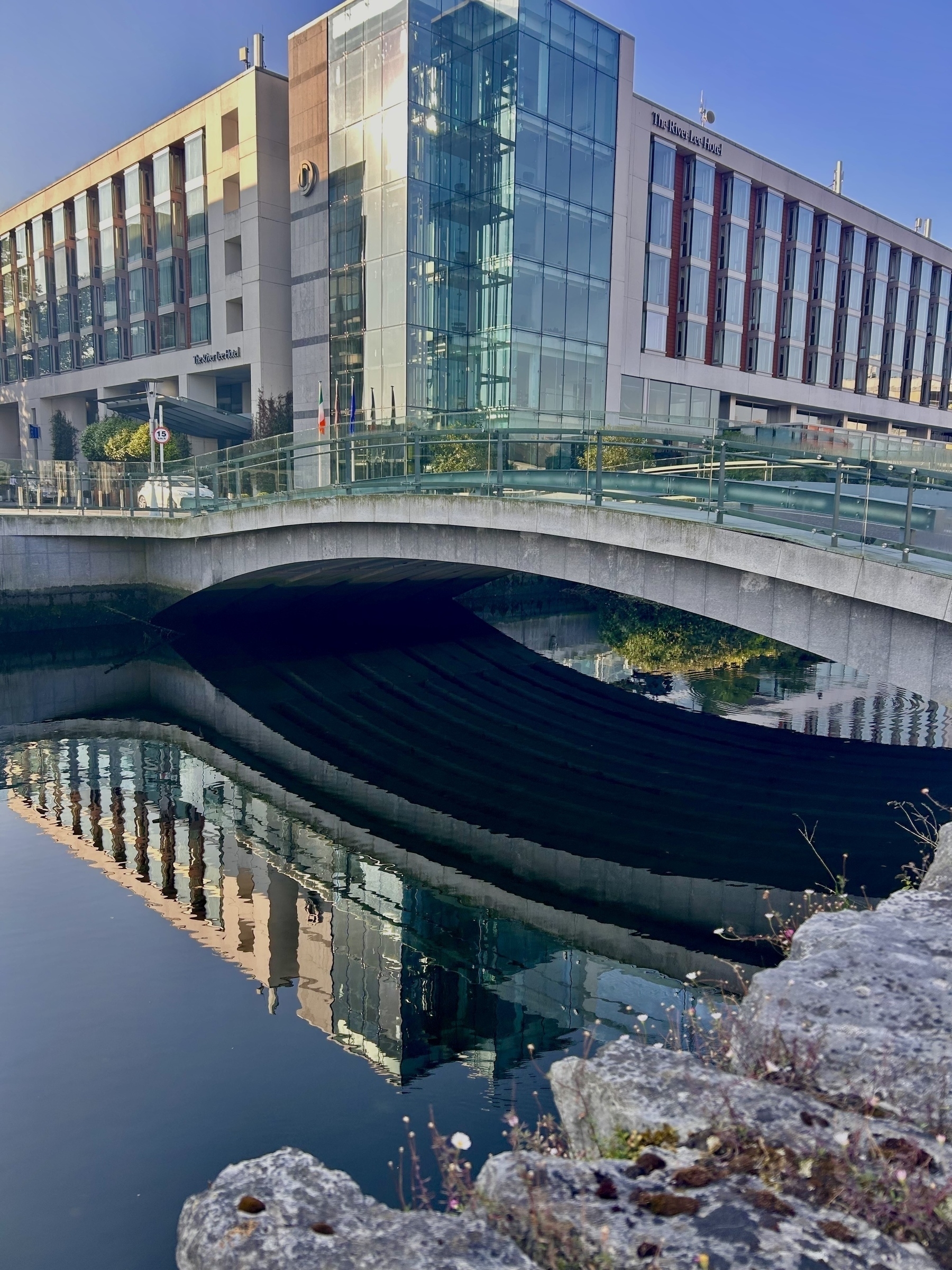 A bridge over the River Lee, with the bridge and a building reflected in the water. 