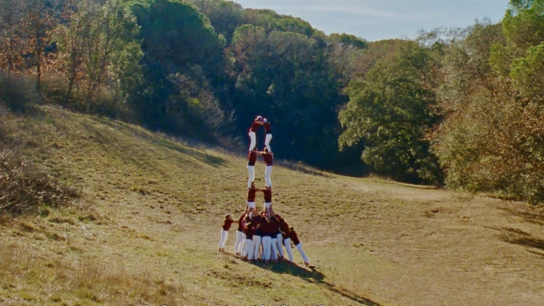 A group of people dressed in red and white on a gently sloping hillside. They are standing on each others’ shoulders to elevate themselves and create a human tower. Fewer on the top, more on the base. It’s a striking image. 