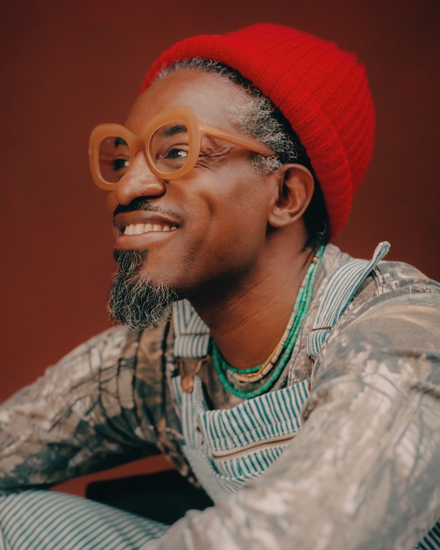 A photograph of André3000, a smiling black man wearing a red beanie and orange glasses, wearing overalls. 