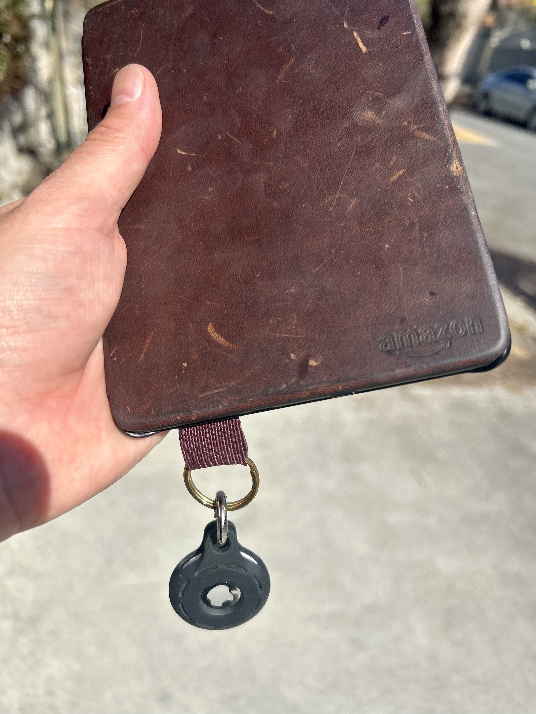 A reader with a brown cover. An AirTag is attached to it using a stuck-on pen loop with a key chain running through it. 