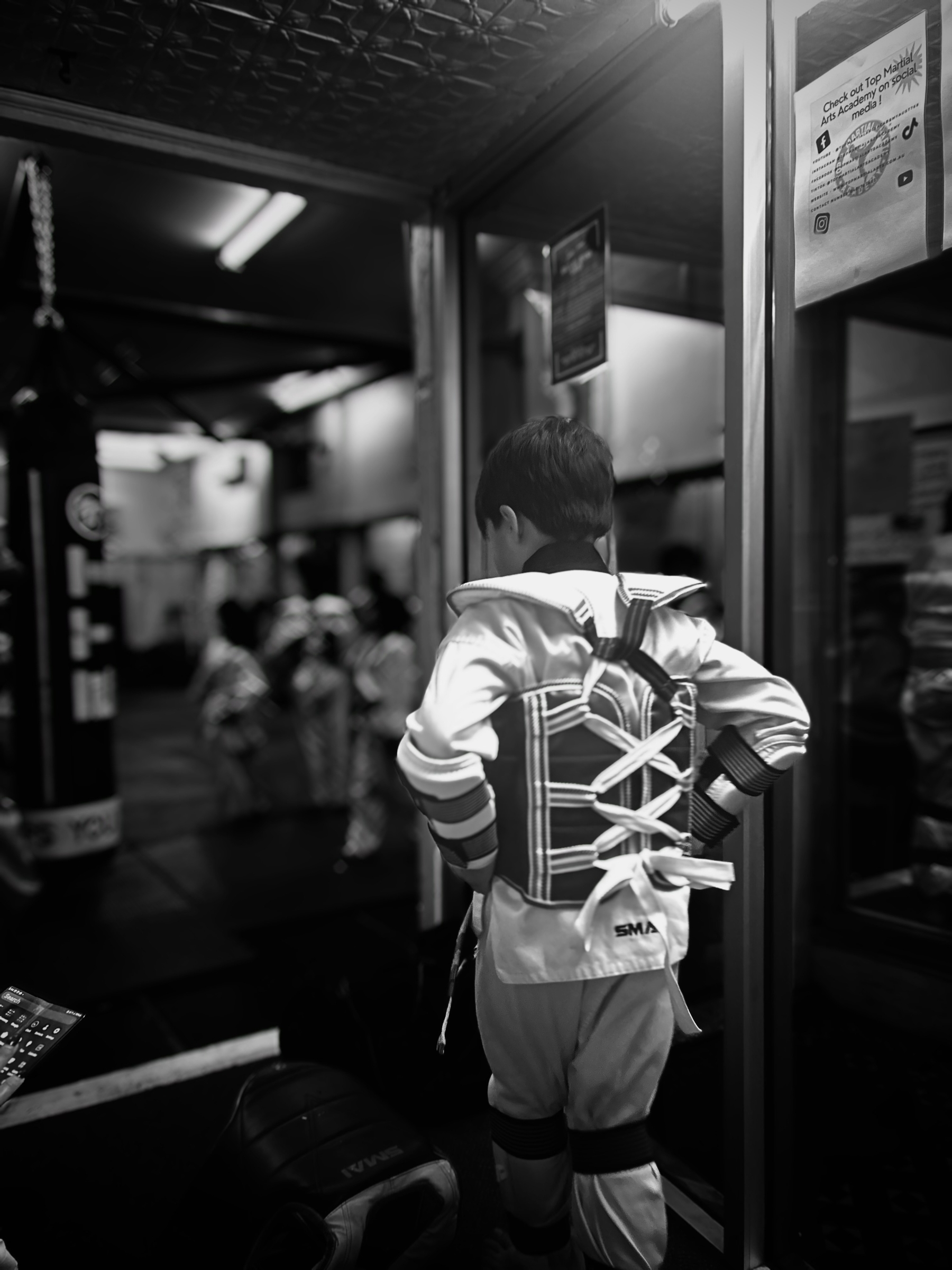 Greyscale image of a youngish child walking into tae kwon do class wearing protective gear 