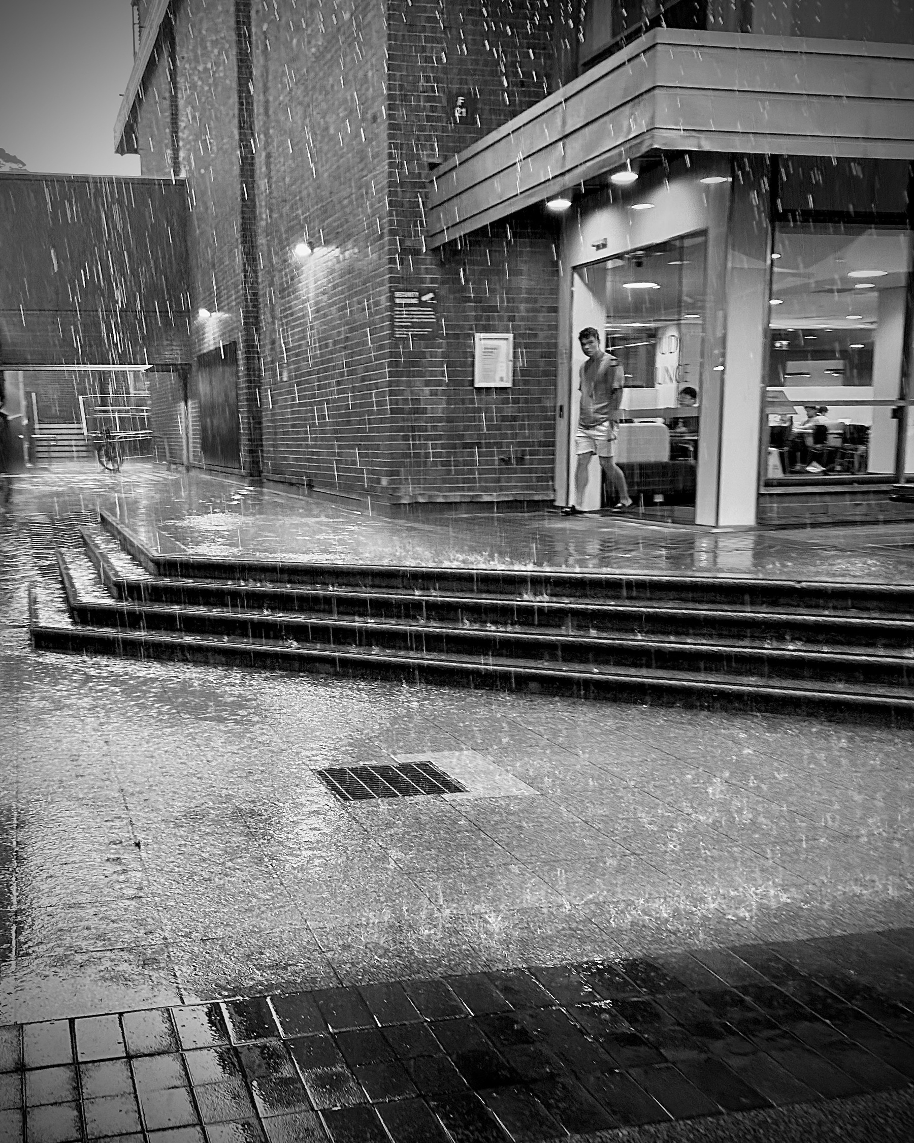 Greyscale image of quite a lot of rain hitting the ground near a drain. In the background someone is exiting a building. 