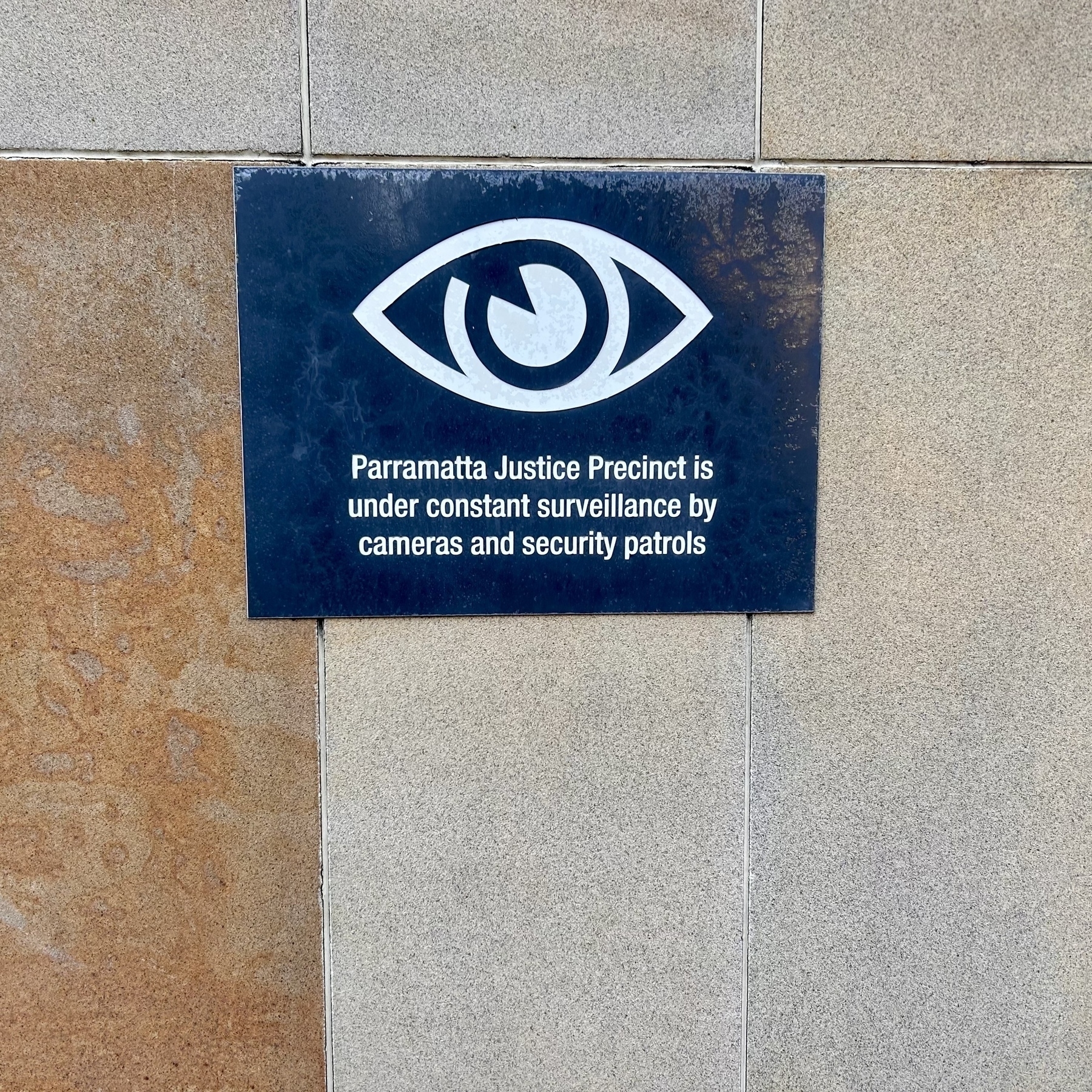 A sign mounted to a wall that says "Parramatta Justice Precinct is under constant surveillance by cameras and security patrols"