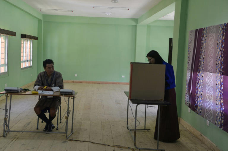 A woman voting at a polling station on Bhutan. A man wearing the traditional gho is seated at a table. 