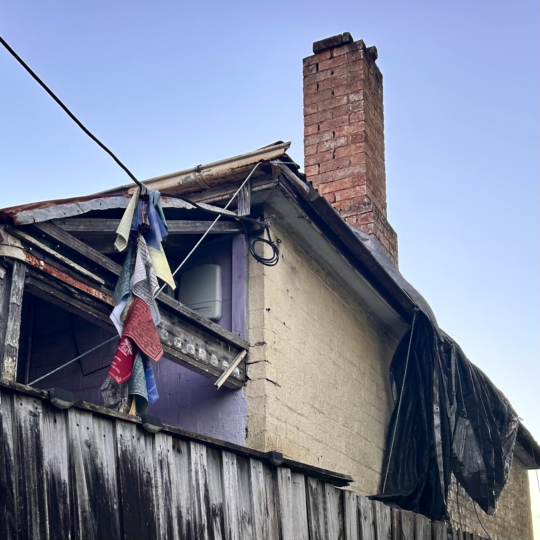 An old house with a tarpaulin hanging off the roof. A faded string of prayer flags is hanging from what I think is the TV cable connection