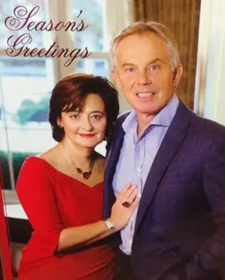 A grimacing couple poses together indoors, with the woman in a red dress and the man in a blue suit, accompanied by the text Season's Greetings  it’s Tony and Cherie Blair.
