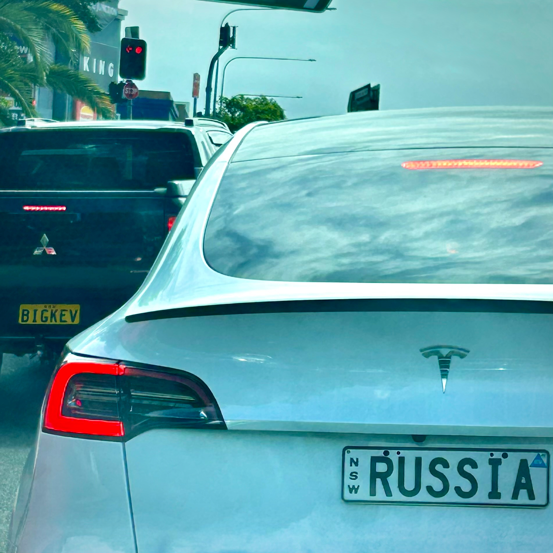 A Tesla with a RUSSIA license plate is stopped at a traffic light next to a black car with the plate BIGKEV.
