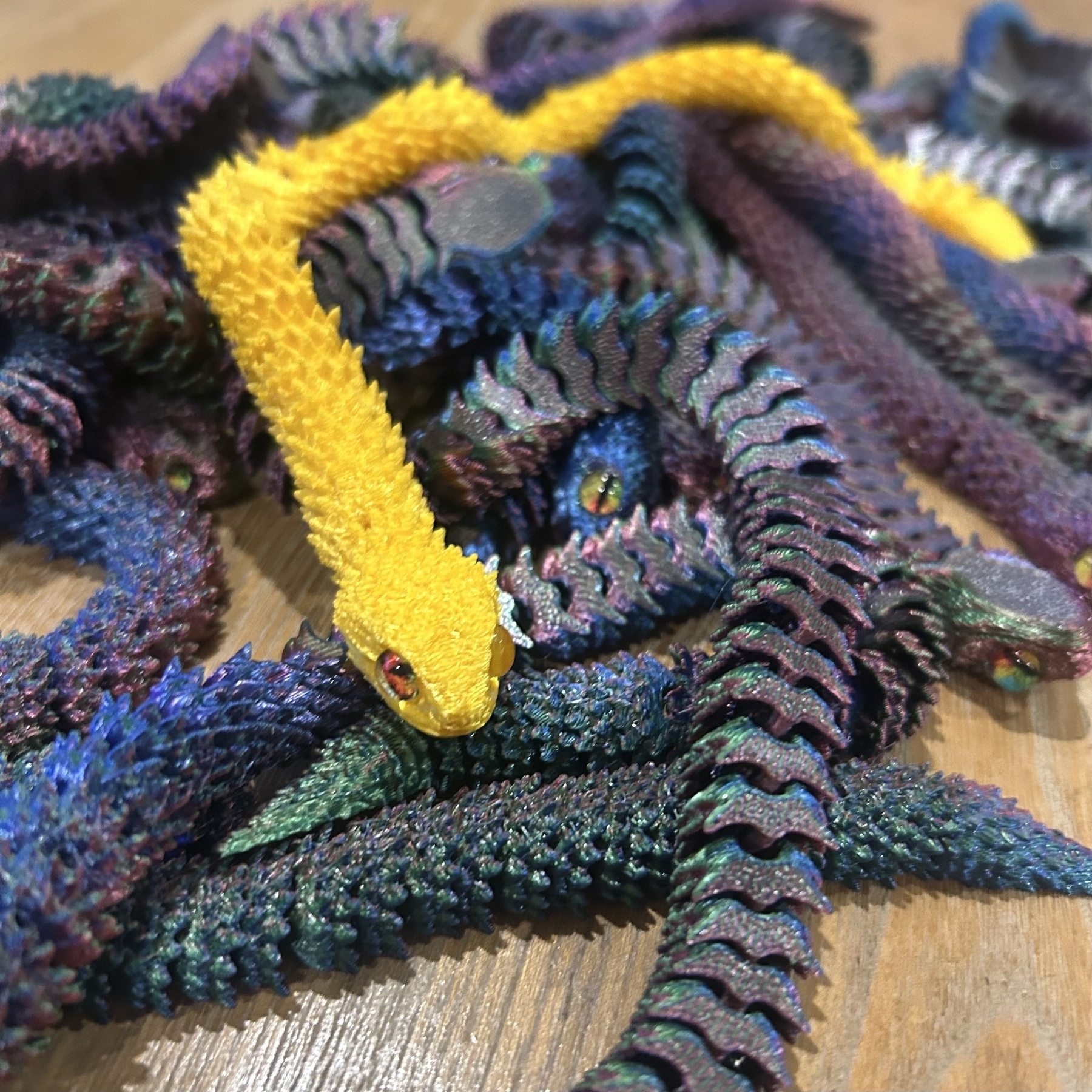 A group of textured, colorful snake-like 3D printed creatures with vibrant scales rests on a wooden surface. The one on top is yellow, being reminiscent of how shiny Pokémon have distinct colours. 