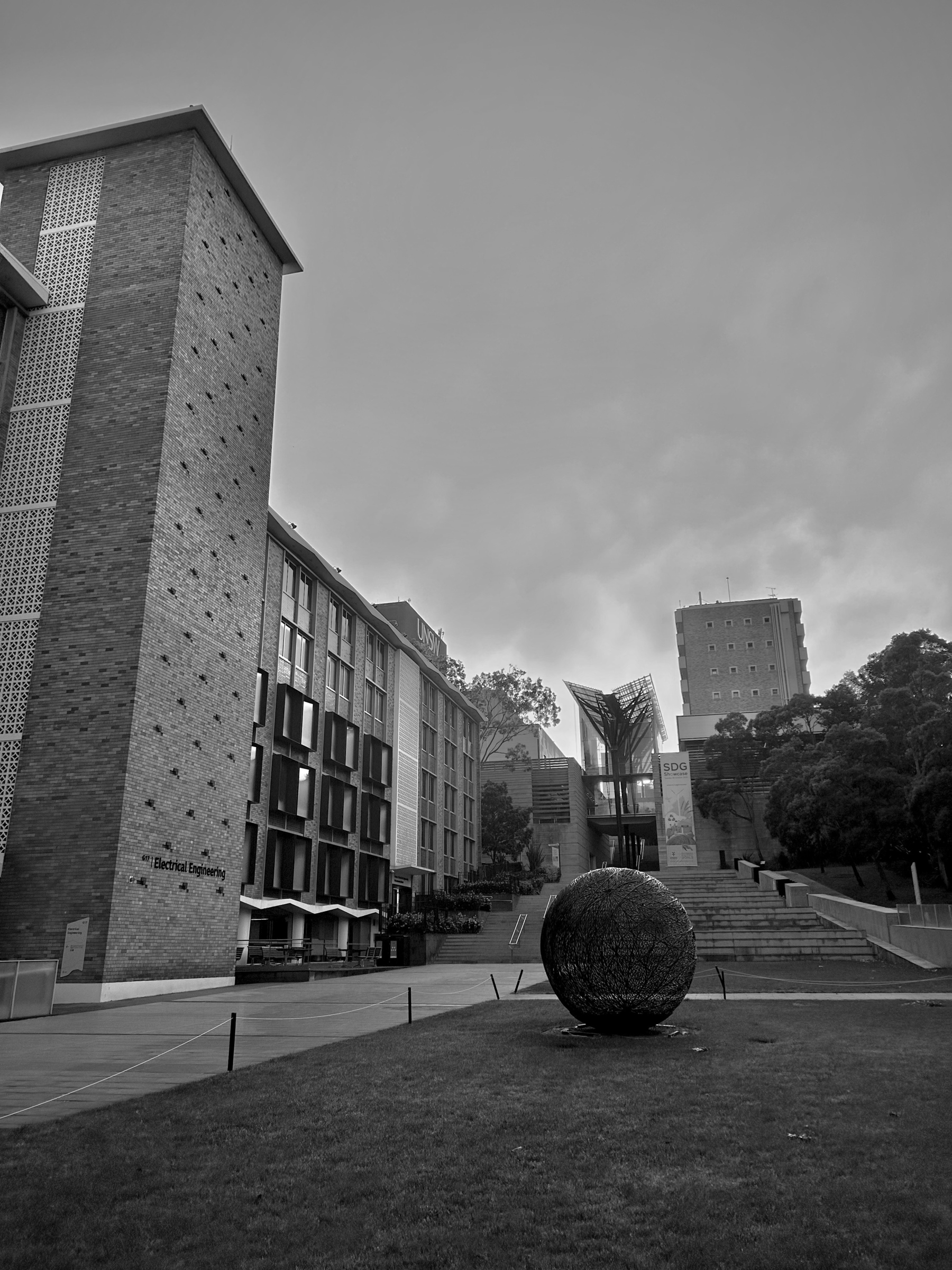UNSW campus in the morning. A modern architectural building complex features a tall brick structure, a large geometric sculpture on the lawn, and a staircase leading to other buildings.