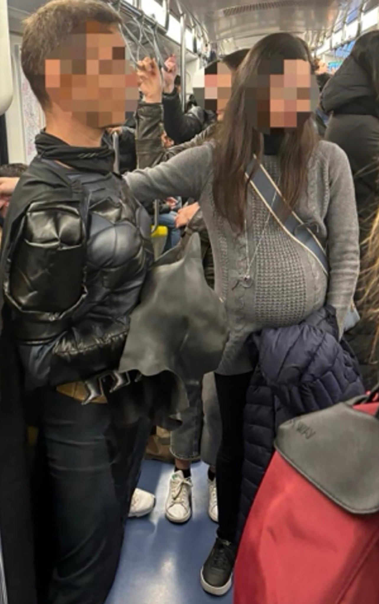 A person dressed as a superhero (Batman costume) stands near a seemingly pregnant person on a crowded subway, surrounded by other standing passengers.