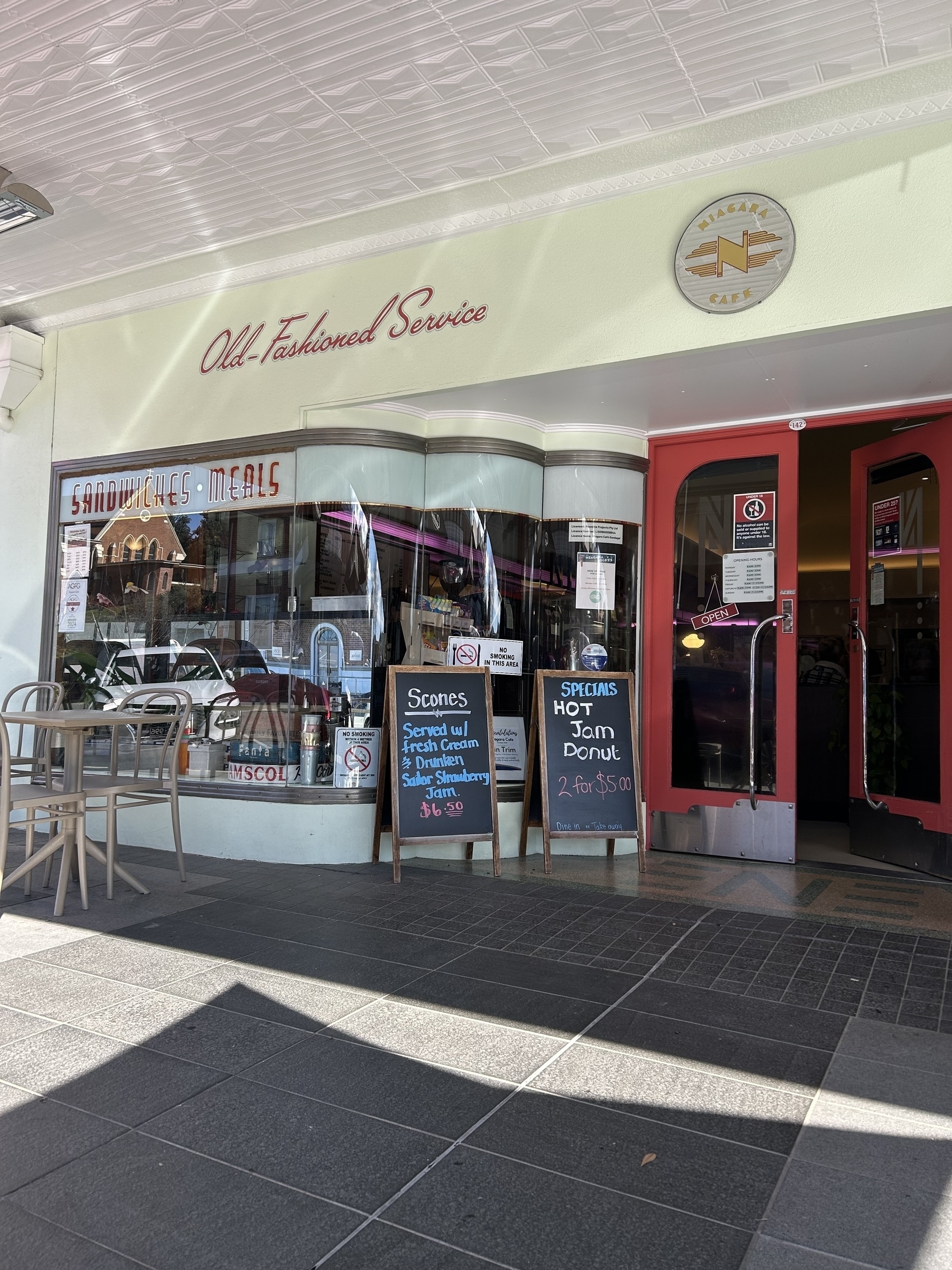 Niagara café in Gundagai with an "Old-Fashioned Service" sign, featuring outdoor seating and chalkboard specials offering scones, jam, and donuts.