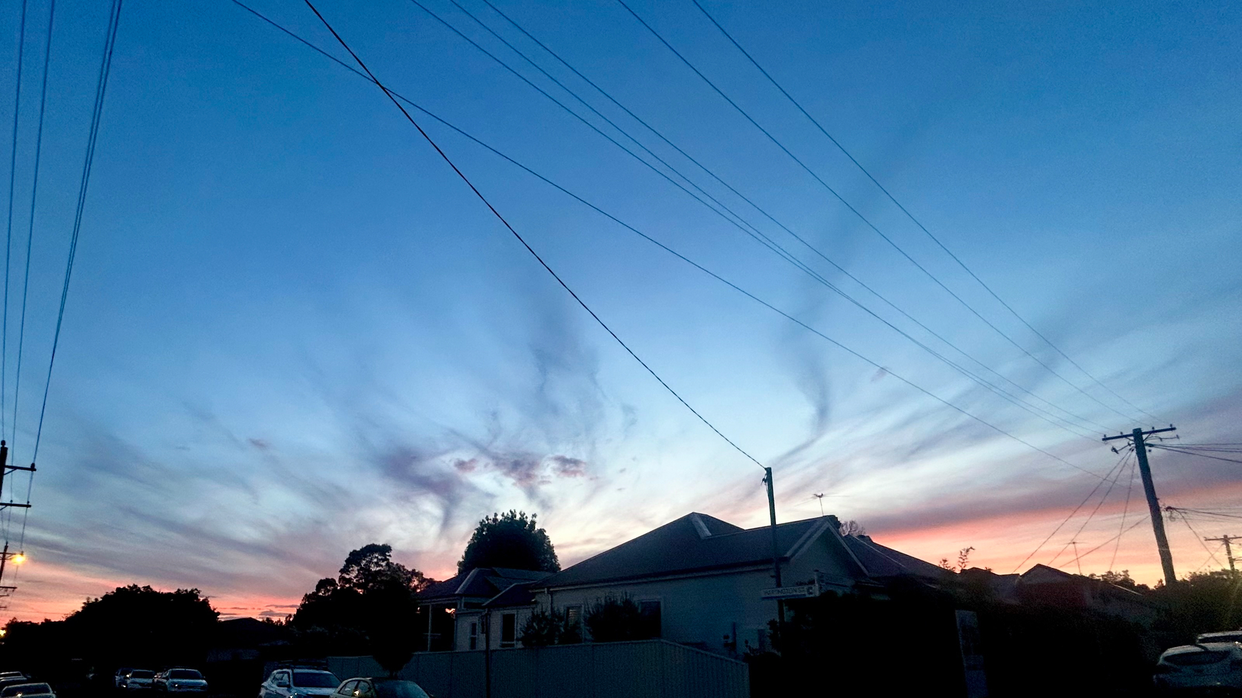 A neighborhood is silhouetted against a colorful evening sky with swirling clouds and power lines overhead.