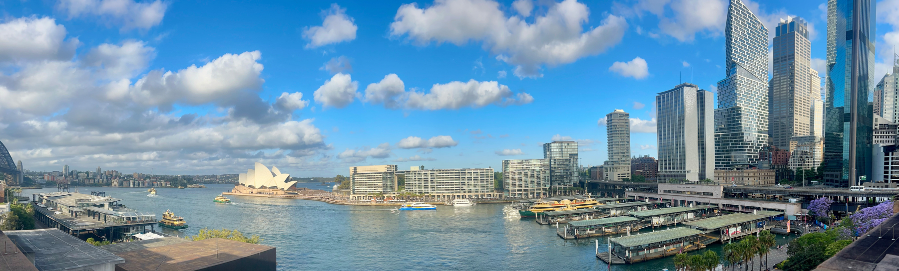 Circular Quay in Sydney. A harbour scene featuring the distinctive Sydney Opera House, ferries, skyscrapers, and a partly cloudy sky.