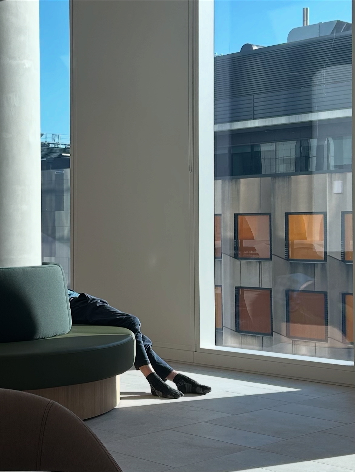A person is relaxing on a round bench by a large window with a view.  They’re not wearing their shoes? 