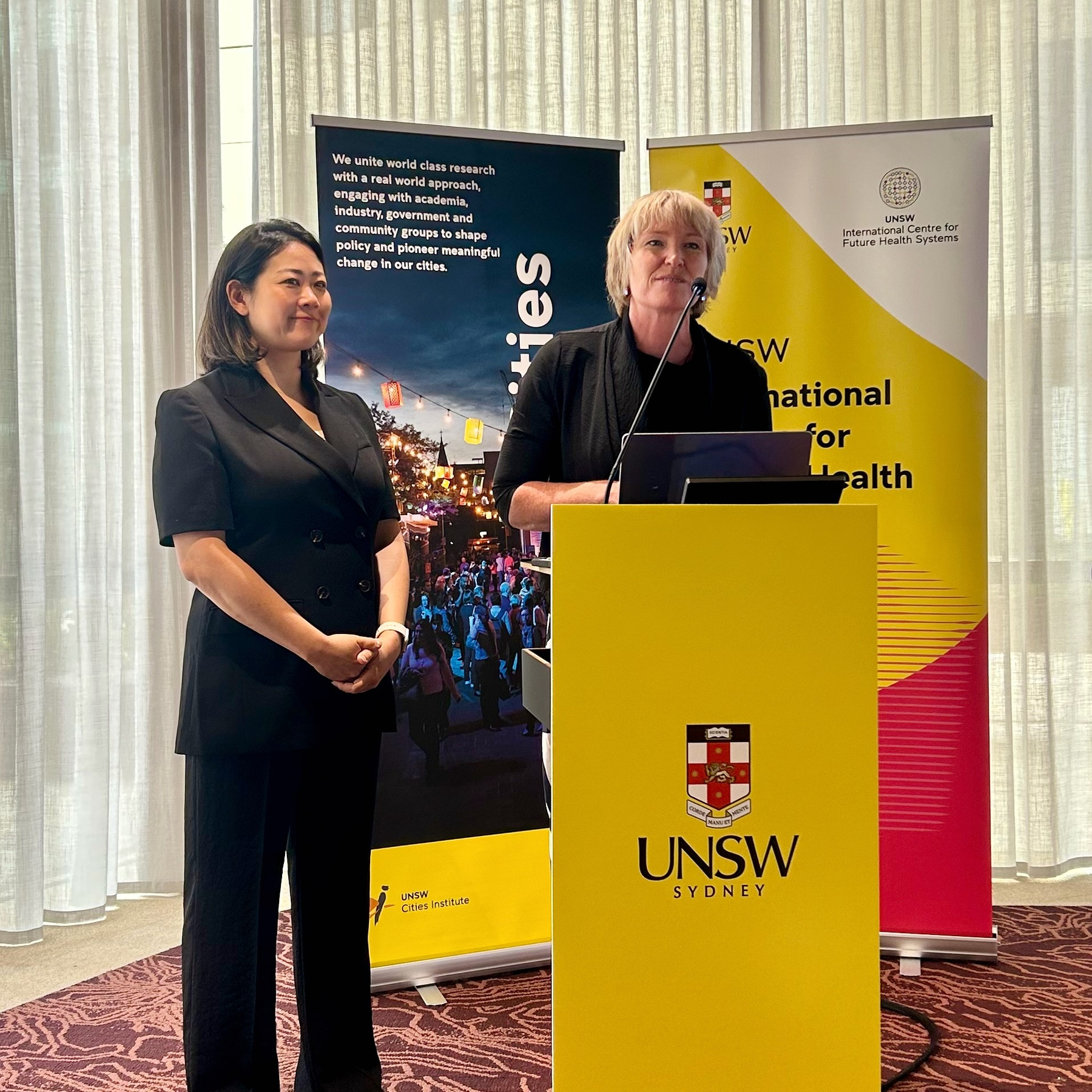 Fiona High and Jinhee Kim stand behind a podium with UNSW Sydney banners in the background, engaged in a presentation.