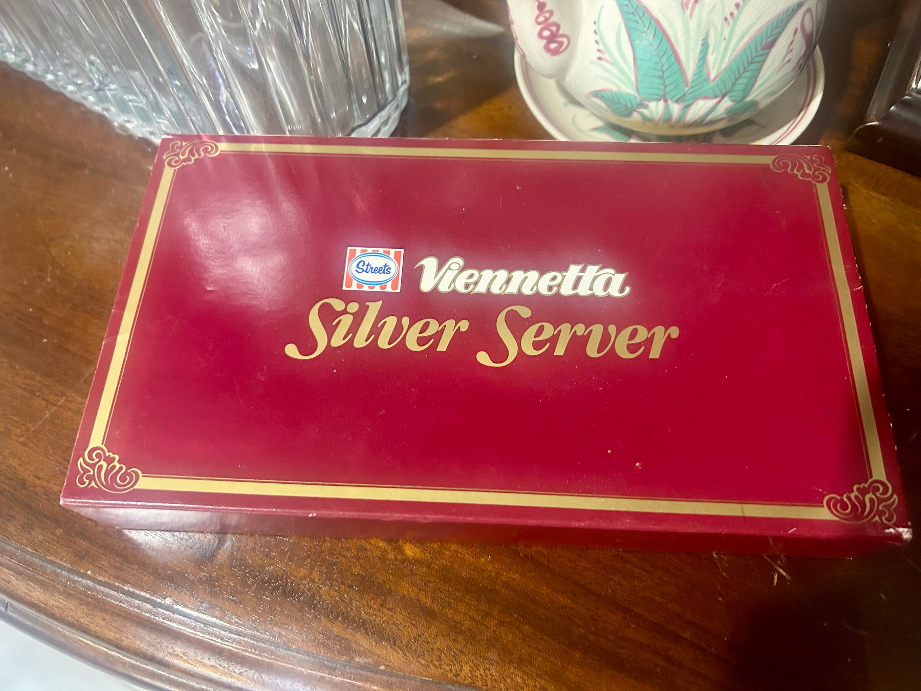 An open red Viennetta Silver Server box sits on a wooden sideboard beside a glass jug and a decorative teacup. Inside the box is a silver-coloured serving spoon designed for serving Viennetta, a layered frozen dessert. It was widely marketed in the ‘80s and ‘90s as a slightly fancy dessert for family dinners and special occasions.