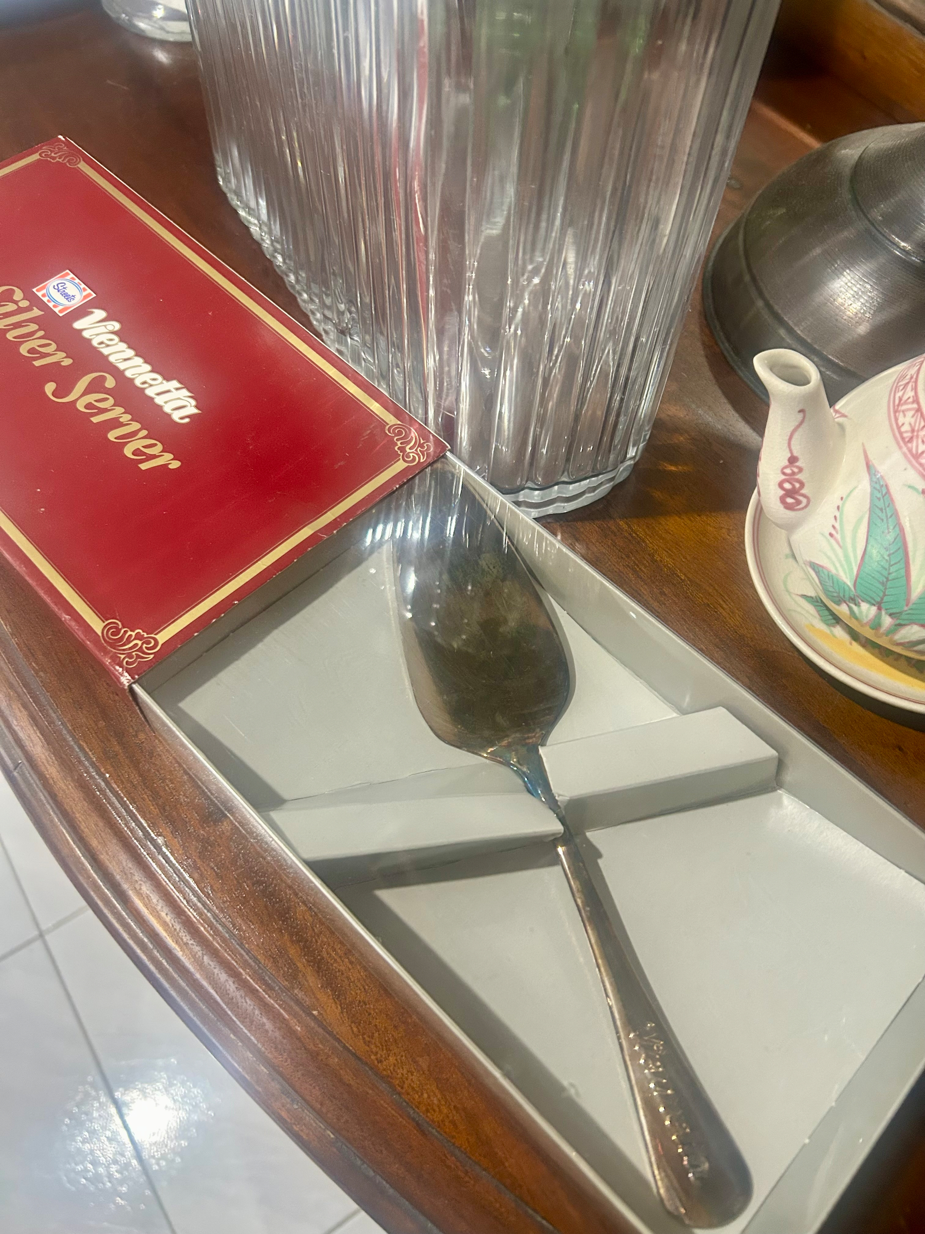 An open red Viennetta Silver Server box sits on a wooden sideboard beside a glass jug and a decorative teacup. Inside the box is a silver-coloured serving spoon designed for serving Viennetta, a layered frozen dessert. It was widely marketed in the ‘80s and ‘90s as a slightly fancy dessert for family dinners and special occasions.