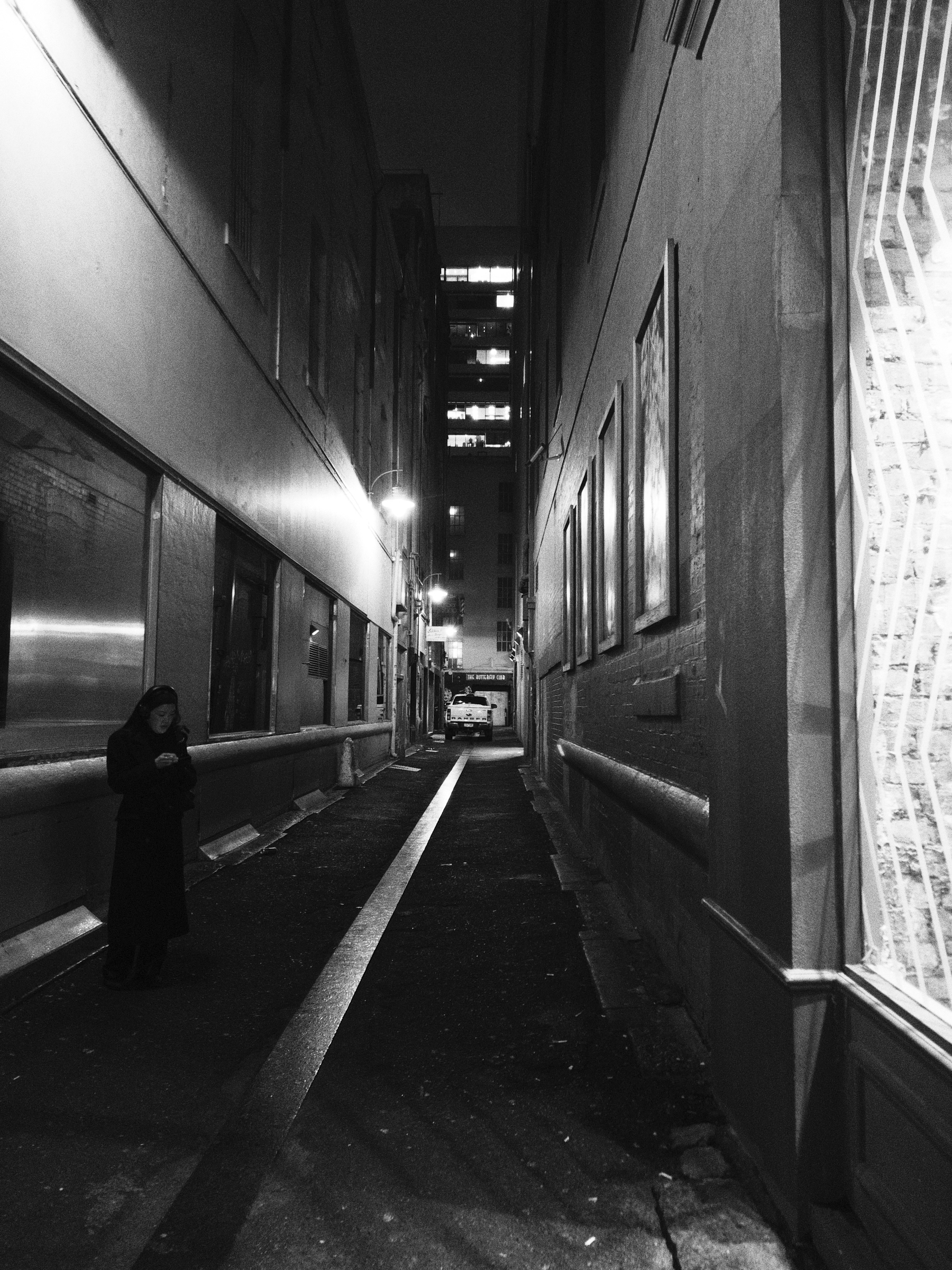 A narrow, dimly lit alleyway at night features a solitary person standing on the left side, with buildings and framed pictures lining the walls.